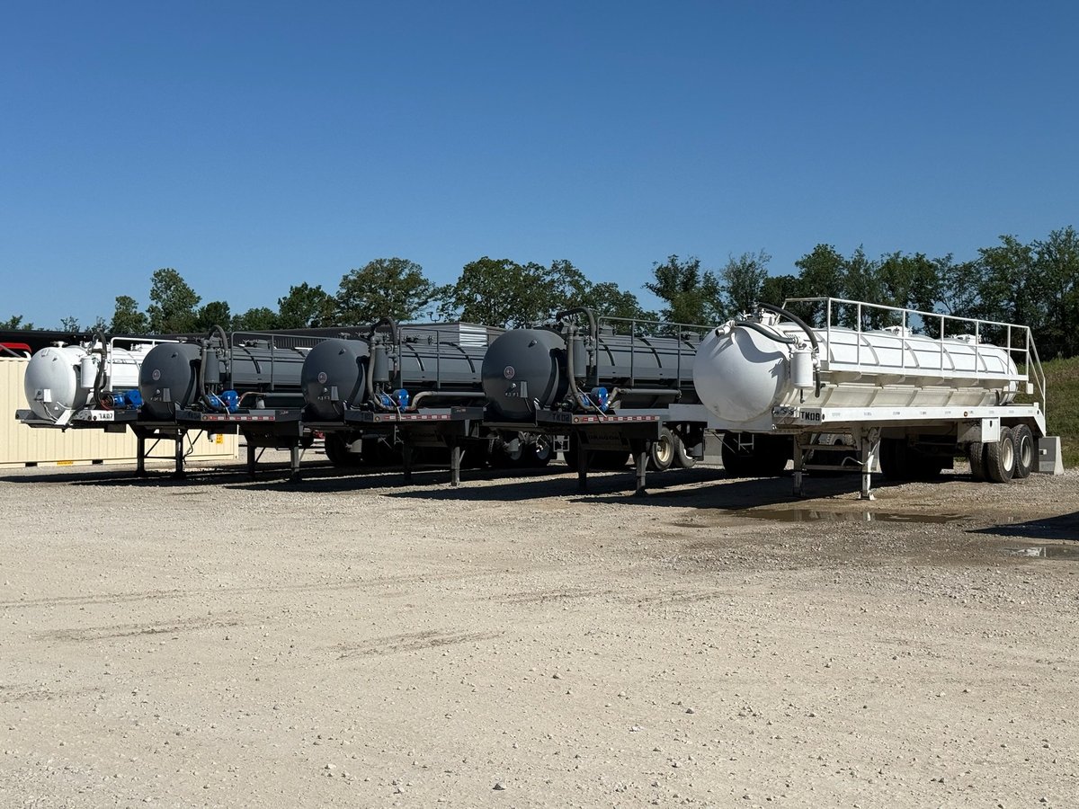 Row of commercial tanker trucks parked on gravel lot with trees and blue sky in background